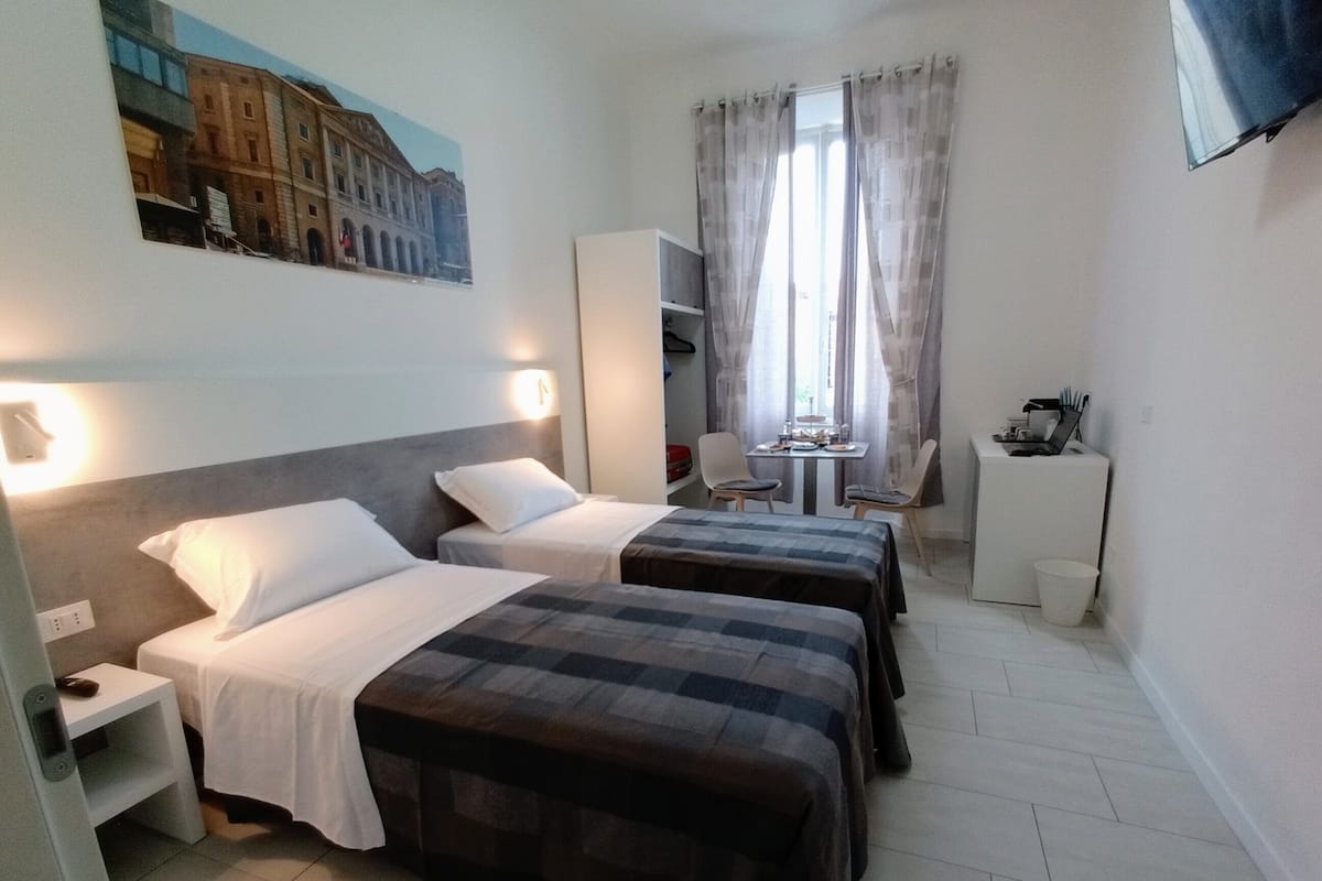 Quarto de hotel com duas camas, uma escrivaninha, uma cadeira, uma mesa pequena e uma janela com cortinas.