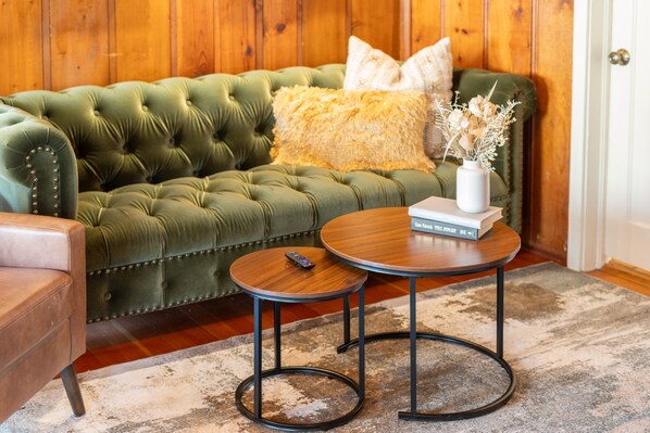 A green tufted sofa with a fluffy pillow, a small round wooden table with a remote control, and a vase with dried flowers. - Pines near Whiskey Row / King Beds / fenced yard (Prescott)