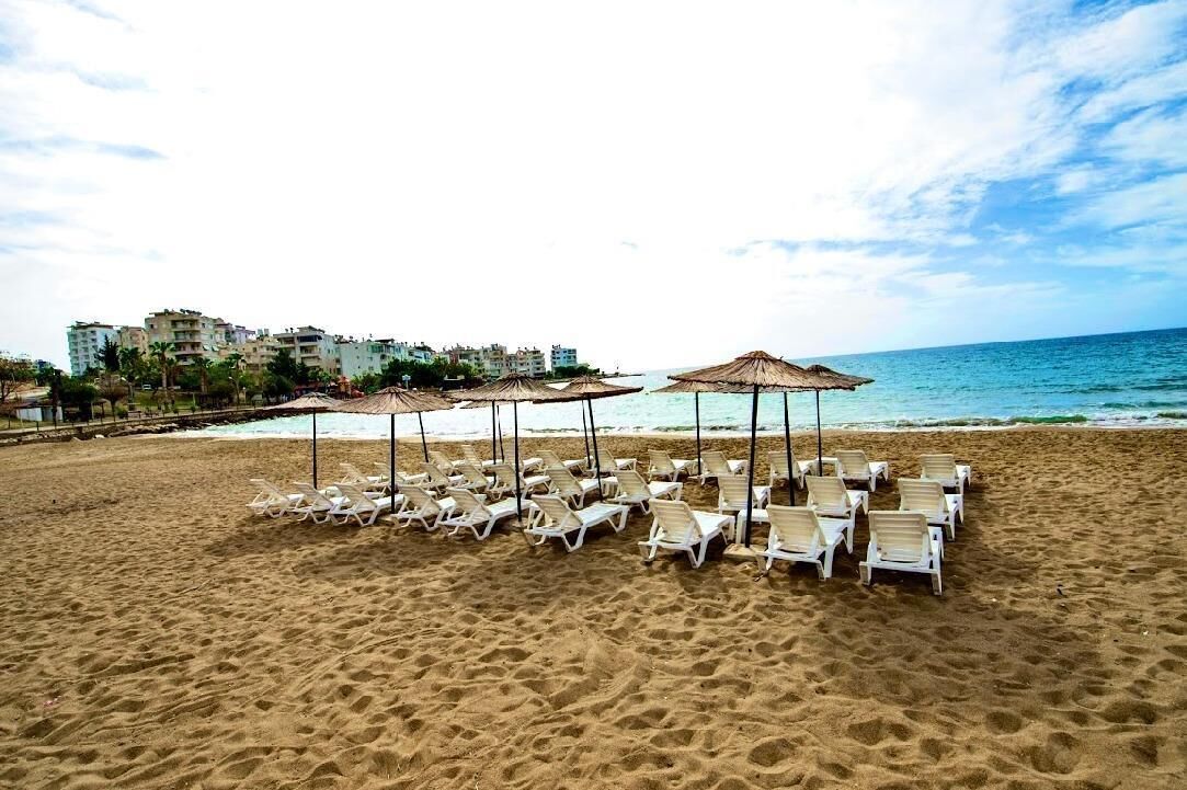Une plage bordée de rangées de chaises longues blanches et d’ombrelles en chaume, une mer calme et des bâtiments en arrière-plan.
