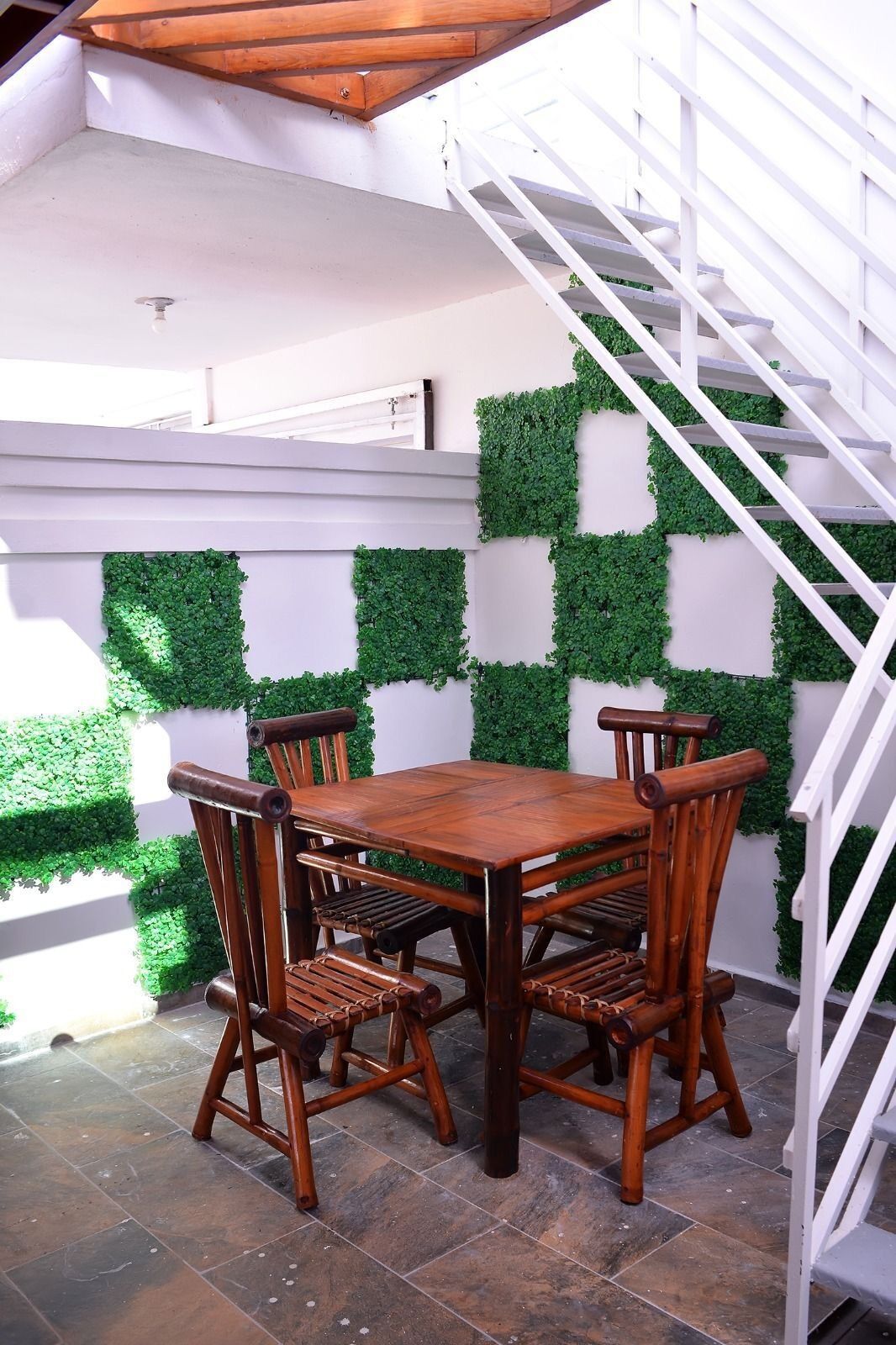 Una mesa de comedor de madera con cuatro sillas, frente a una pared blanca con follaje verde. Una escalera blanca es visible a la derecha.