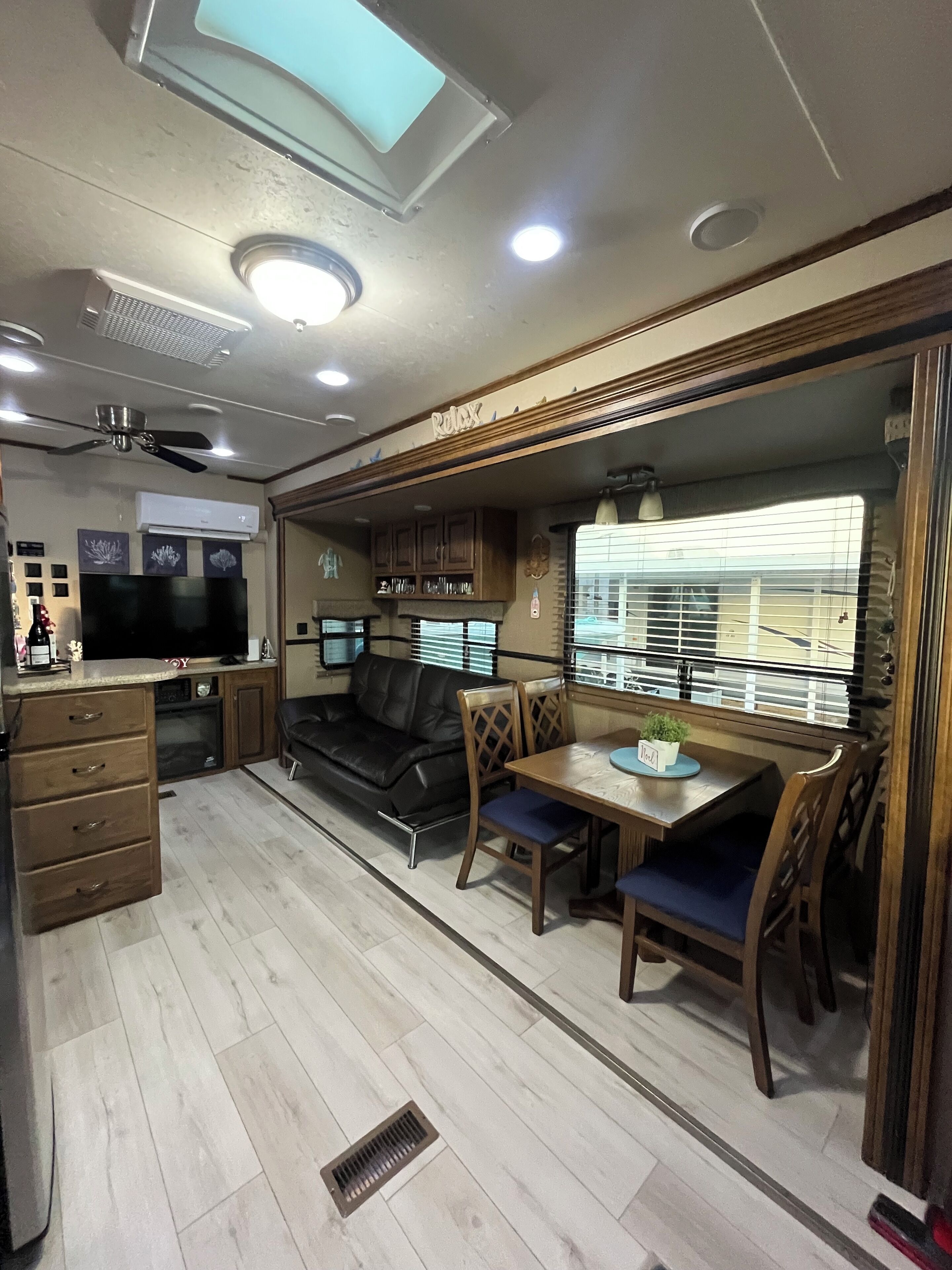 A compact RV interior with a kitchenette, a dining area, a seating area with a black leather couch, and a ceiling fan.