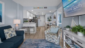 Una sala de estar con sofá, sillón y mesa de centro, una zona de cocina y una mesa de comedor con sillas.