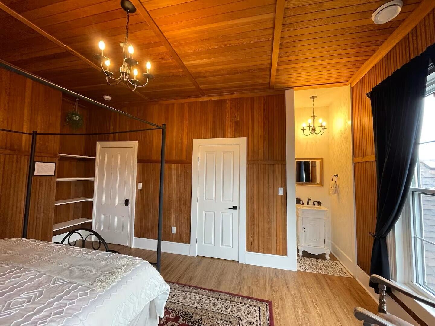 A bedroom with wooden paneling, a bed with a patterned bedspread, a chandelier, a bathroom with a mirror, and a window with curtains.