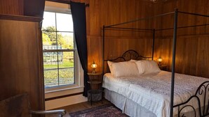 A four-poster bed with a white bedspread, a wooden chair, a small table with a lamp, and a view of a garden through a window.