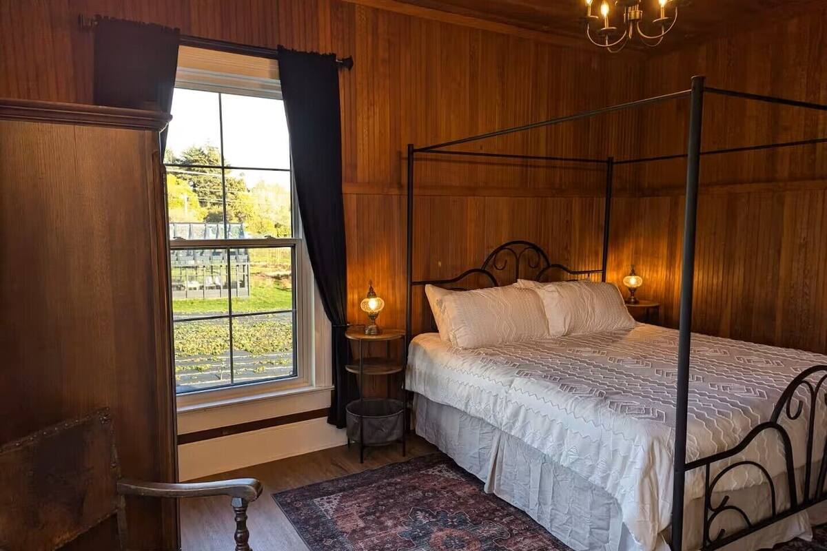 A four-poster bed with a white bedspread, a wooden chair, a small table with a lamp, and a view of a garden through a window.