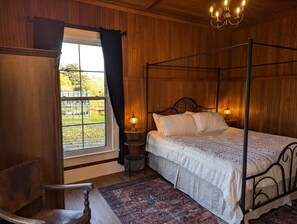 A four-poster bed with a white bedspread, a wooden chair, a small table with a lamp, and a view of a garden through a window.