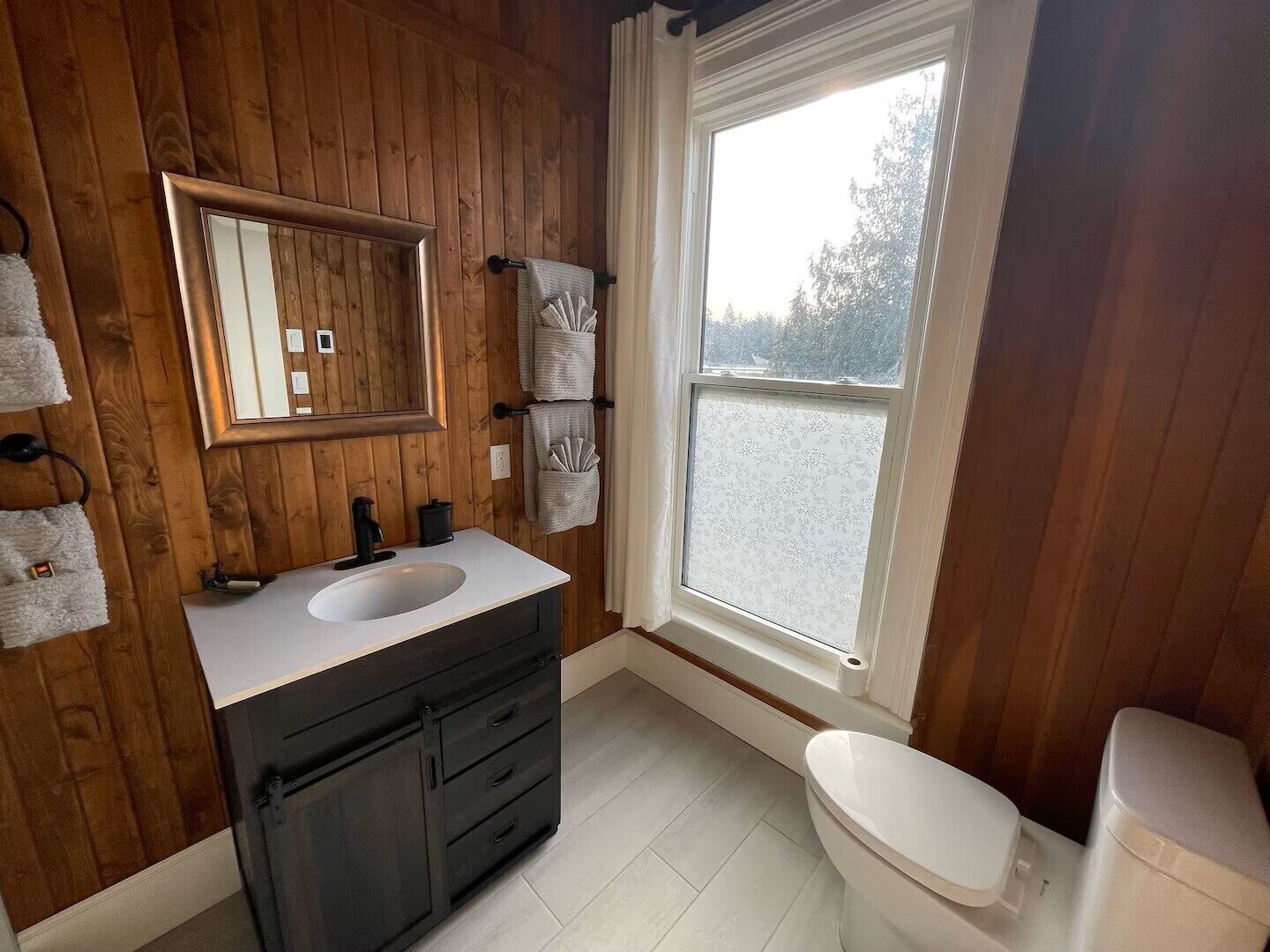 A bathroom with wooden walls, a white sink, black cabinets, a toilet, and a window with a view.