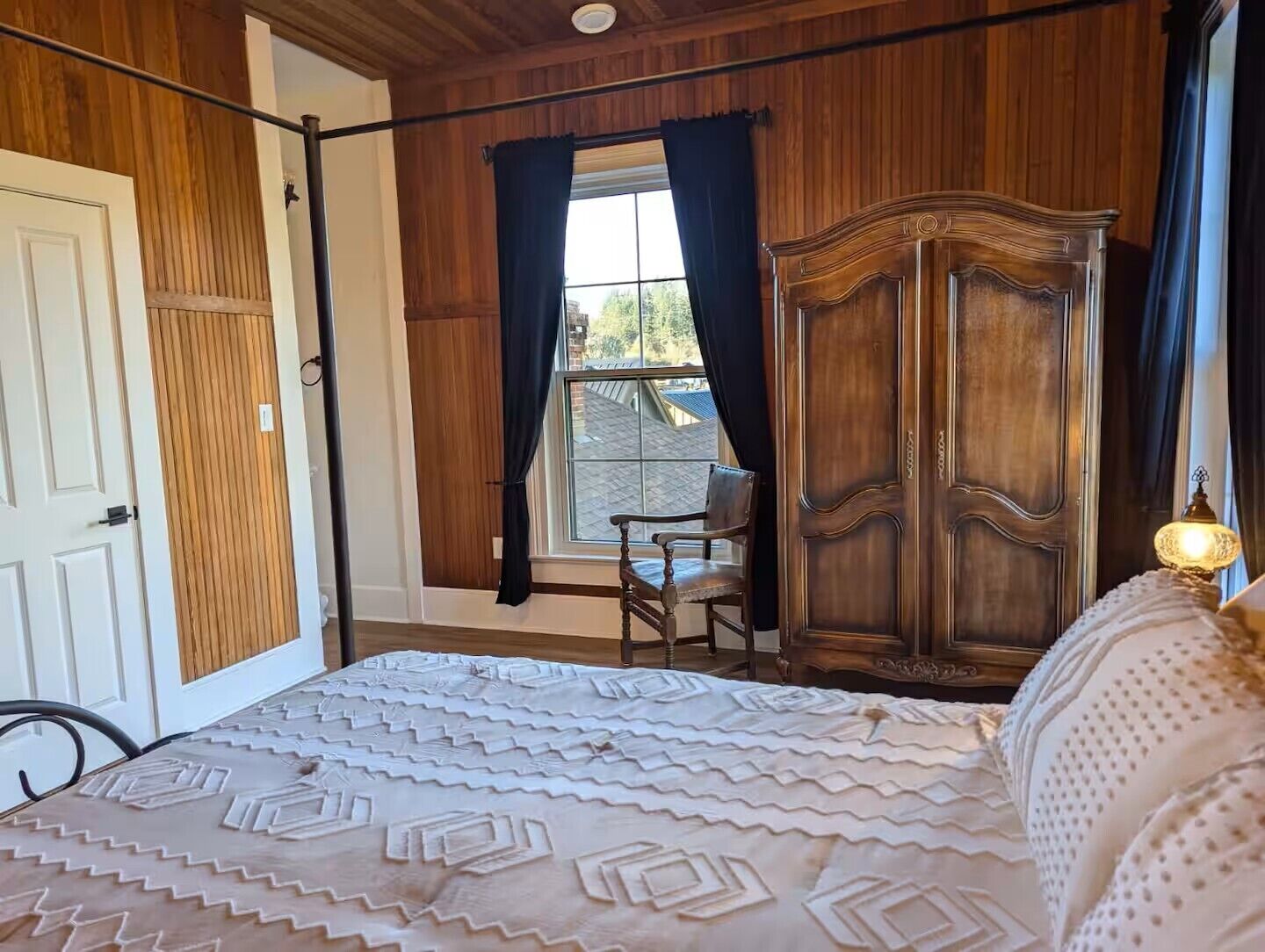 A bedroom with a wooden wardrobe, a bed with a patterned bedspread, a chair, and a window with curtains.