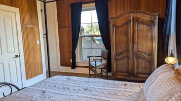 A bedroom with a wooden wardrobe, a bed with a patterned bedspread, a chair, and a window with curtains.