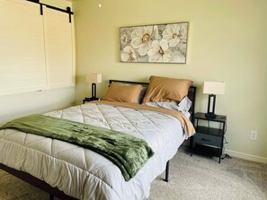 Ein ordentlich bezogenes Bett mit einer grünen Tagesdecke, zwei Nachttischen mit Lampen und einem floralen Kunstwerk an der Wand.