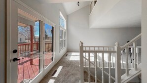 Una habitación amplia con un gran ventanal corredero que da a una terraza exterior, una escalera blanca y un techo alto con tragaluces.