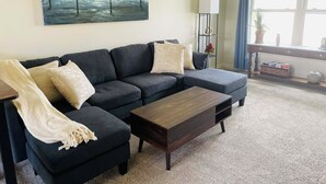 Una sala de estar con un sofá seccional gris oscuro, una mesa de centro de madera y un cuadro con árboles en la pared.
