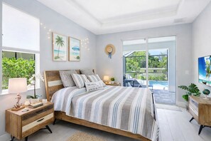 A bedroom with a bed, bedside tables, a nightstand, a window with blinds, a wall-mounted hat, a framed picture, and a view of a pool and greenery outside.