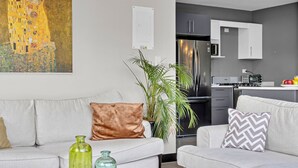 Een moderne woonkamer met twee banken, een salontafel met boeken en decoratieve flessen, een groot schilderij aan de muur, en een keukenhoek met een koelkast en kasten.