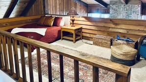 Ein gemütliches Schlafzimmer im Dachgeschoss mit Holzboden, einem Ziegelkamin, einem hölzernen Bett mit roter Bettwäsche und einem kleinen Tisch mit Lampe.