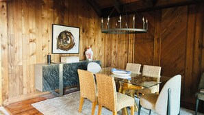 Ein Holzinterieur mit einem Esstisch, Korbstühlen, einem Tisch mit Glasplatte, einer Holzbalkendecke und einer Kandelaber.