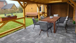 A covered patio with a wooden table and chairs, a bench, and a flower planter. - Simones-Ferienhaus.de Cozy vacation in North Friesland for up to 6 people. (Drage)