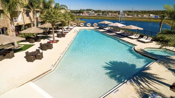 Ein großer rechteckiger Pool mit Liegestühlen und Sonnenschirmen, umgeben von Palmen und Blick auf ein weiter entferntes Gewässer.
