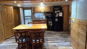 Een houten interieur met een eettafel en stoelen, een keuken met een koelkast en fornuis, en een raam met uitzicht op bomen.
