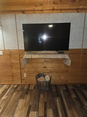 Ein Zimmer mit an der Holzwand befestigtem Fernseher, einem gewebten Regal darunter und einem Korb mit aufgerollten Teppichen auf dem Boden.