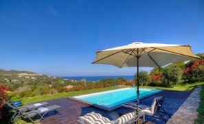 Ein Swimmingpool mit Liegestühlen und Sonnenschirm, umgeben von Grün und Blick aufs Meer.