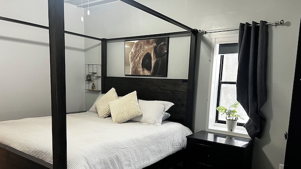 A four-poster bed with a dark headboard, a bedside table with a plant, and a window with curtains. - HUGE Gameroom-Style Barndo in the countryside, minutes to Lake McMurtry & OSU! (Stillwater)