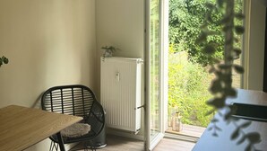 A dining area with a wooden table and a black chair, a white radiator, and a window with a view of greenery. - Beautiful apartment in a quiet location with good connections to Potsdam and Berlin (Potsdam)
