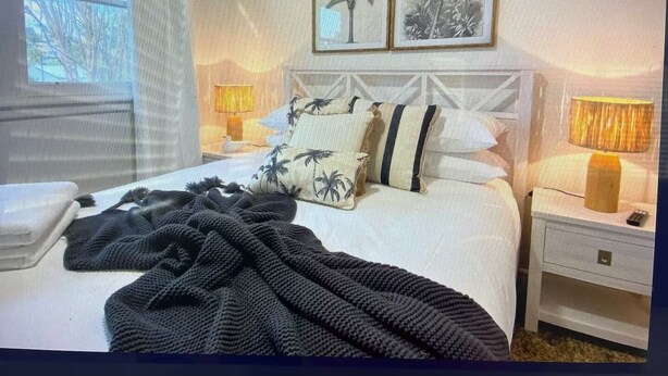 Een net opgemaakt bed met een zwart-wit gestreept dekbed, twee kussens met palmboommotieven, en een hoofdbord met een geometrisch patroon. Er staan twee nachtlampen met lampenkappen, een witte nachtkastje, en een raam met zonwering.