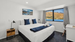 Habitación de hotel con cama, escritorio, silla, mesita de noche y ventana con vista a montañas y un lago.