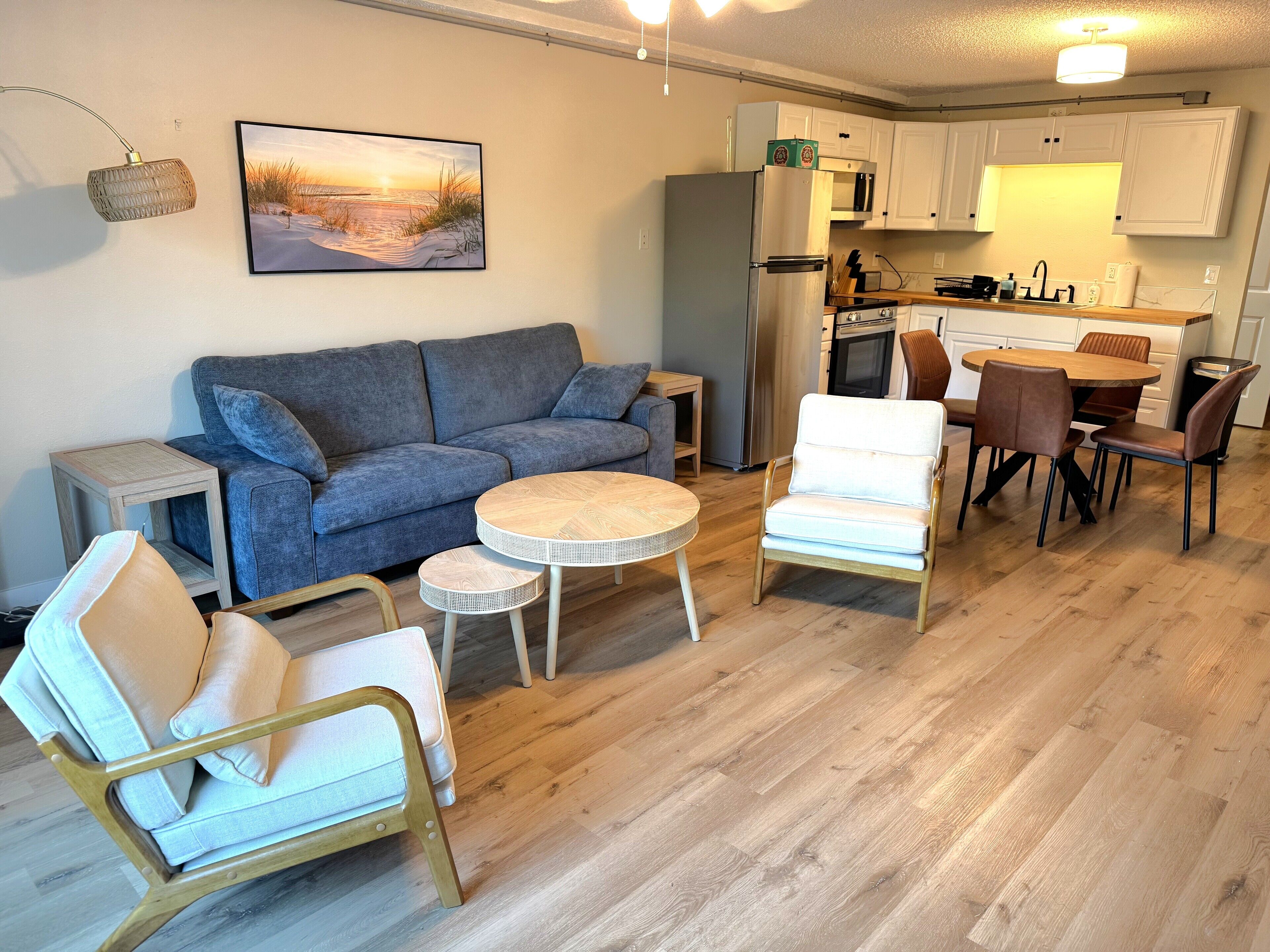 Un salon moderne avec un canapé gris, une table basse ronde en bois et un fauteuil blanc. On aperçoit, au fond, une cuisine avec des meubles blancs et une table à manger en bois.