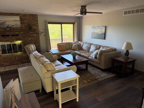 A living room with a sofa, armchair, coffee table, and a lamp. There is a fireplace, a window with a view, and a ceiling fan.