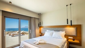 A hotel room with a large bed, a nightstand, a wooden headboard, and a view of the sea through a sliding glass door. - Anemelia Villas (Karpathos)