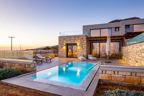A modern outdoor pool area with stone walls, lounge chairs, and a glass balustrade. - Anemelia Villas (Karpathos)