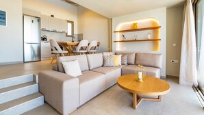 A modern living room with a sectional sofa, a round coffee table, and a dining area with chairs and a table. - Anemelia Villas (Karpathos)
