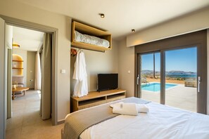 A bedroom with a bed, a TV, a wooden shelf, a robe, and a view of a swimming pool and landscape. - Anemelia Villas (Karpathos)