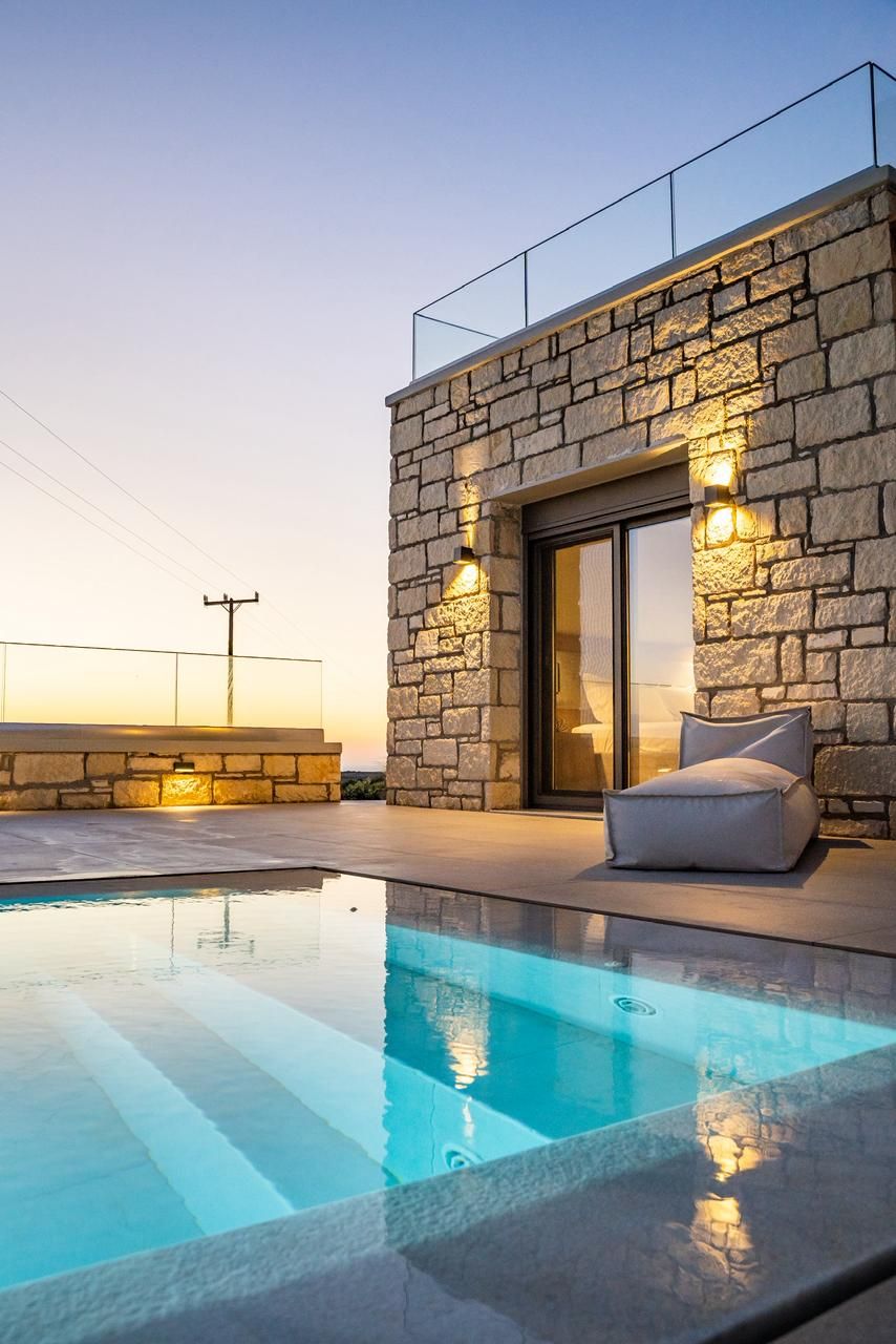 Ein moderner Poolbereich mit Steinwand und Glastüren, einer gepolsterten Sitzgruppe und freiem Blick zum Himmel.