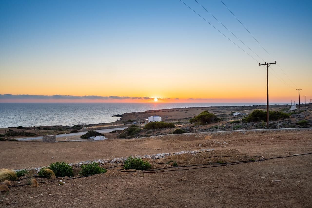 A coastal landscape with a sunset, a dirt path, sparse vegetation, and a few buildings.