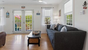 A living room with a dark gray sofa, a wooden coffee table, and a ceiling fan. There are two doors leading to a balcony with an American flag. - Walk to the Waves: Play & Unwind on the Coast! (Waveland)