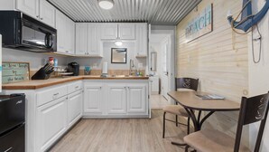 A kitchen with white cabinets, a black microwave, and a wooden countertop. There is a sign on the wall that reads "BEACH". - Walk to the Waves: Play & Unwind on the Coast! (Waveland)