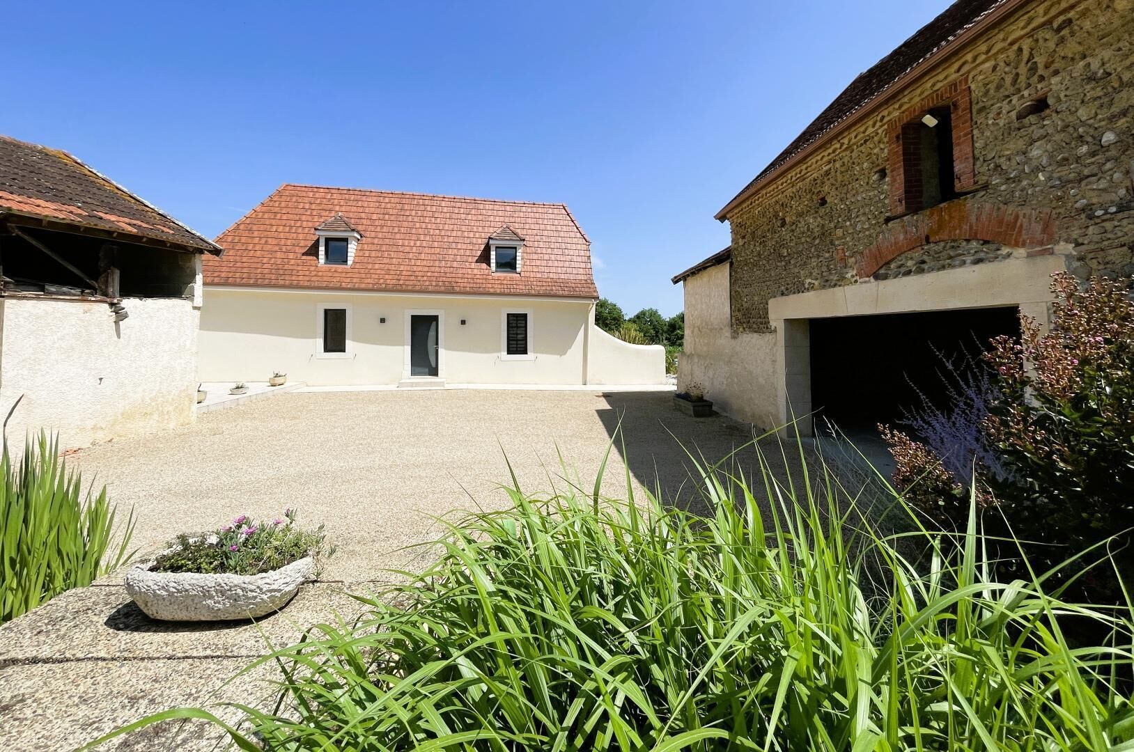 A courtyard with a white building featuring a red-tiled roof, a stone building with a garage, and a stone planter with plants.