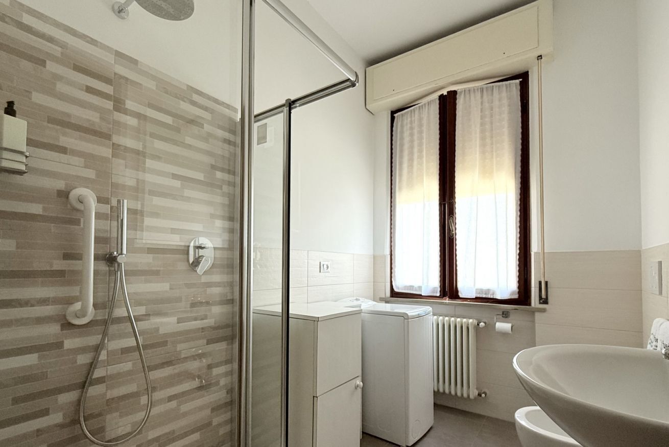 A modern bathroom with a glass shower enclosure, a built-in washing machine, a radiator, and a sink.