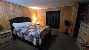Un dormitorio con una pared revestida de madera, una cama con colcha, una lámpara de noche, un aparador y un espejo.