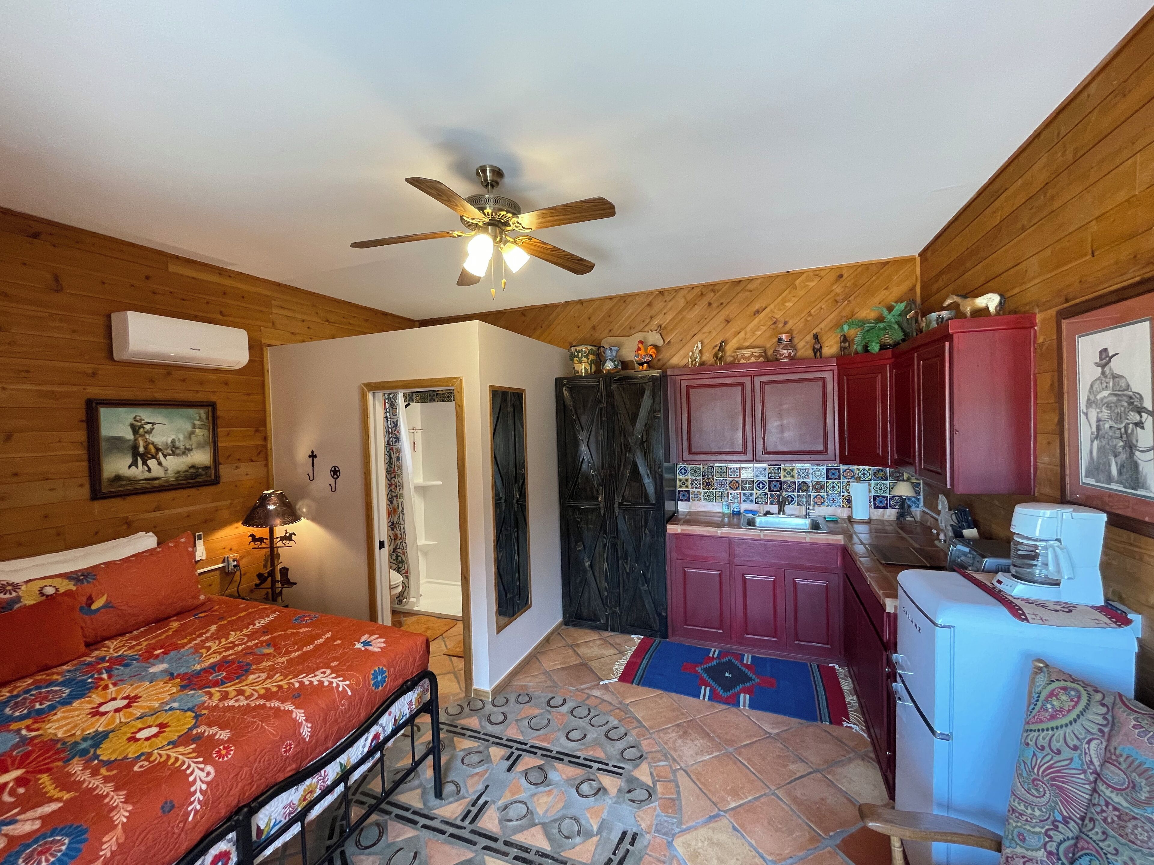 A compact room with a bed, a small kitchenette, a ceiling fan, and a door leading to another room.