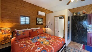 Um quarto com teto de madeira, cama com colcha florida, ventilador de teto, janela com persianas e porta que dá para outro cômodo.