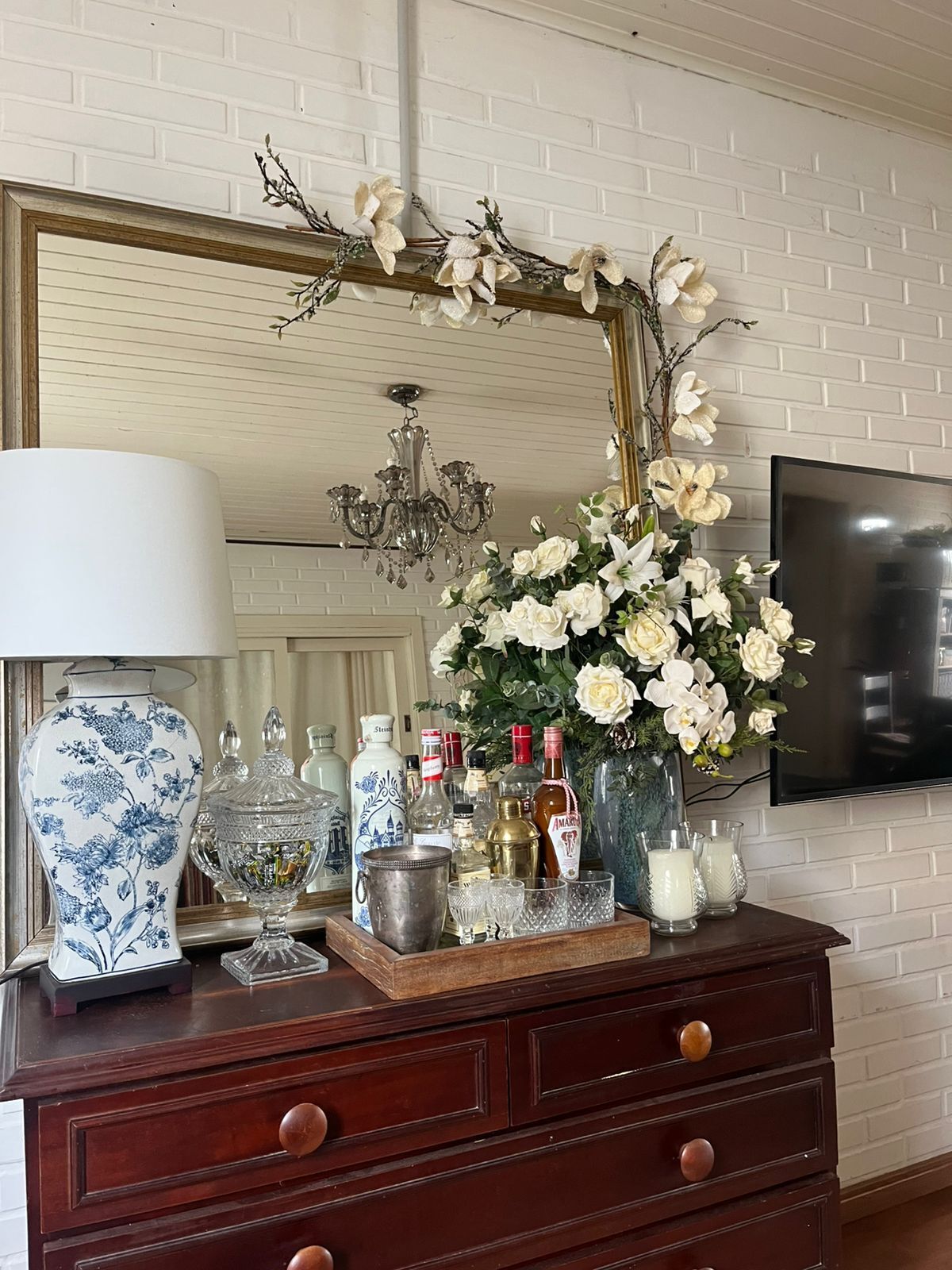 A wooden dresser with a mirror, a vase of white flowers, and various bottles and glasses.