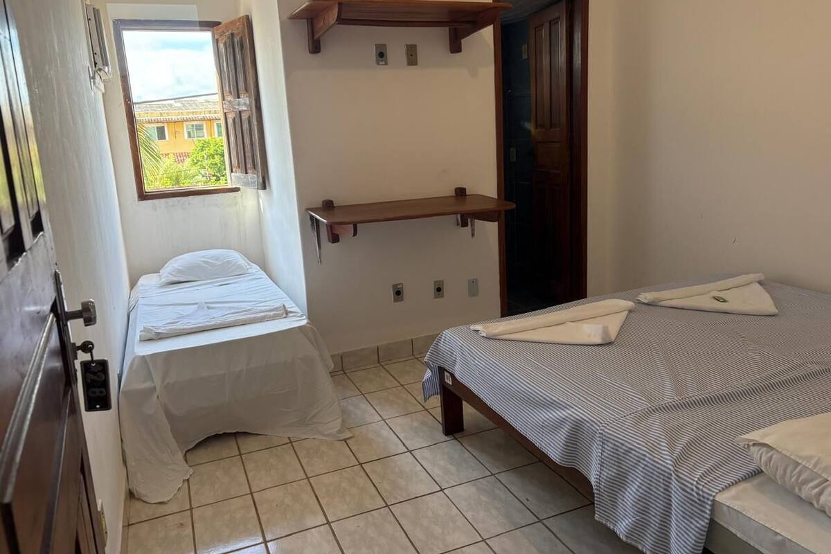 A small, clean room with two beds, a window, and a wooden shelf.