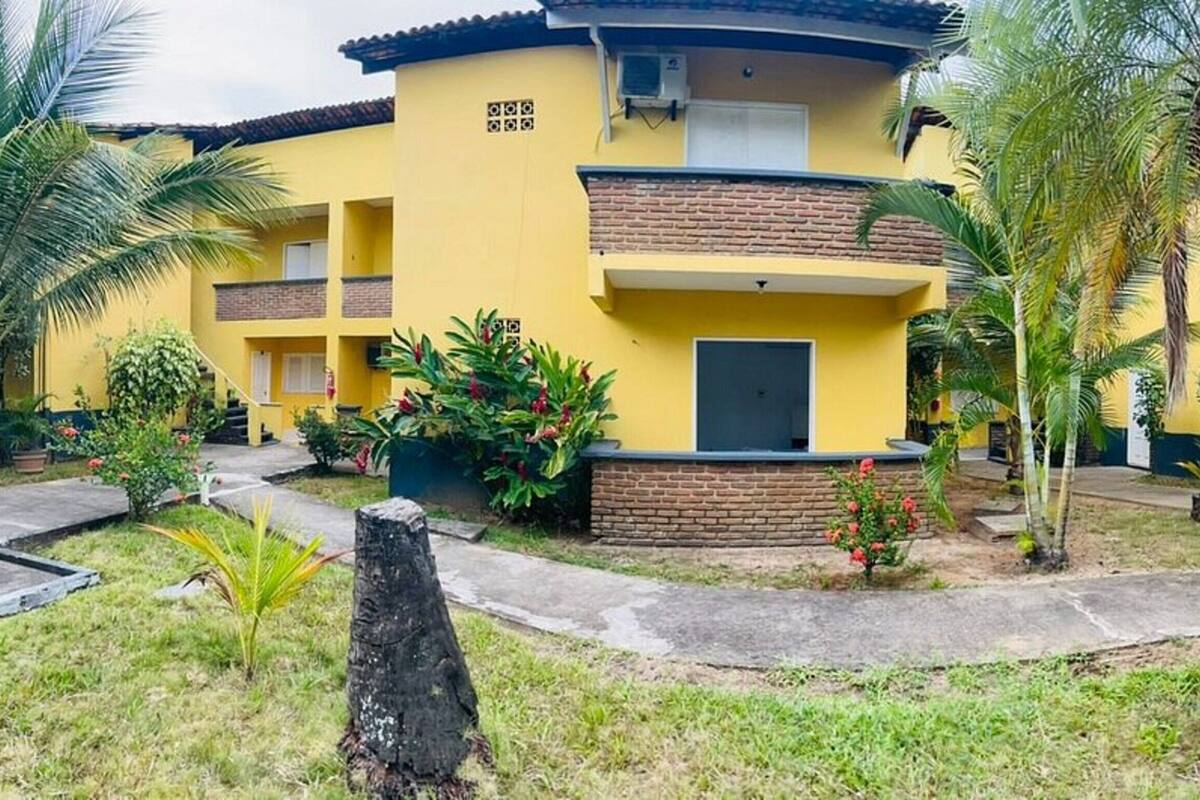 A yellow building with a brick base and a balcony, surrounded by palm trees and small plants.