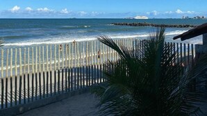 Una playa con una cerca de madera, palmeras y vista al océano y edificios lejanos.