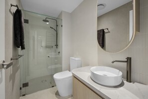 A modern bathroom with a glass-enclosed shower, a white toilet, a sink with a marble countertop, and a mirror with a round frame. - Bilinga Beachfront Luxury at Lana (Bilinga)