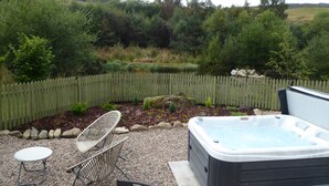 A hot tub in a garden with a wooden fence, surrounded by rocks and plants. - Shepherds  View -  a tiny home that sleeps 2 guests  in 1 bedroom (Glenshee)